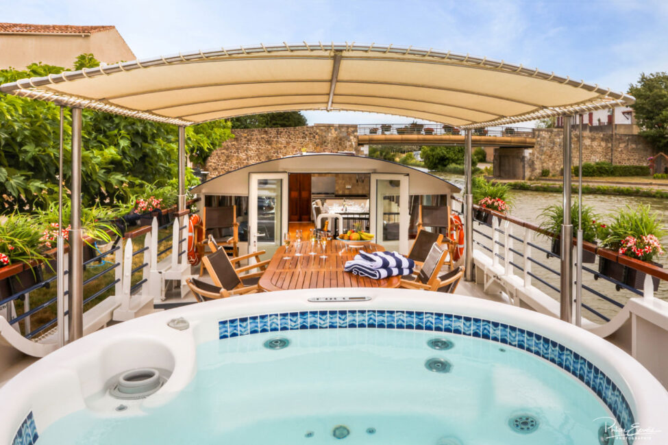 Jacuzzi sur le pont d'une péniche avec en arrière plan une table en bois, des verres de champagne et un espace restaurant