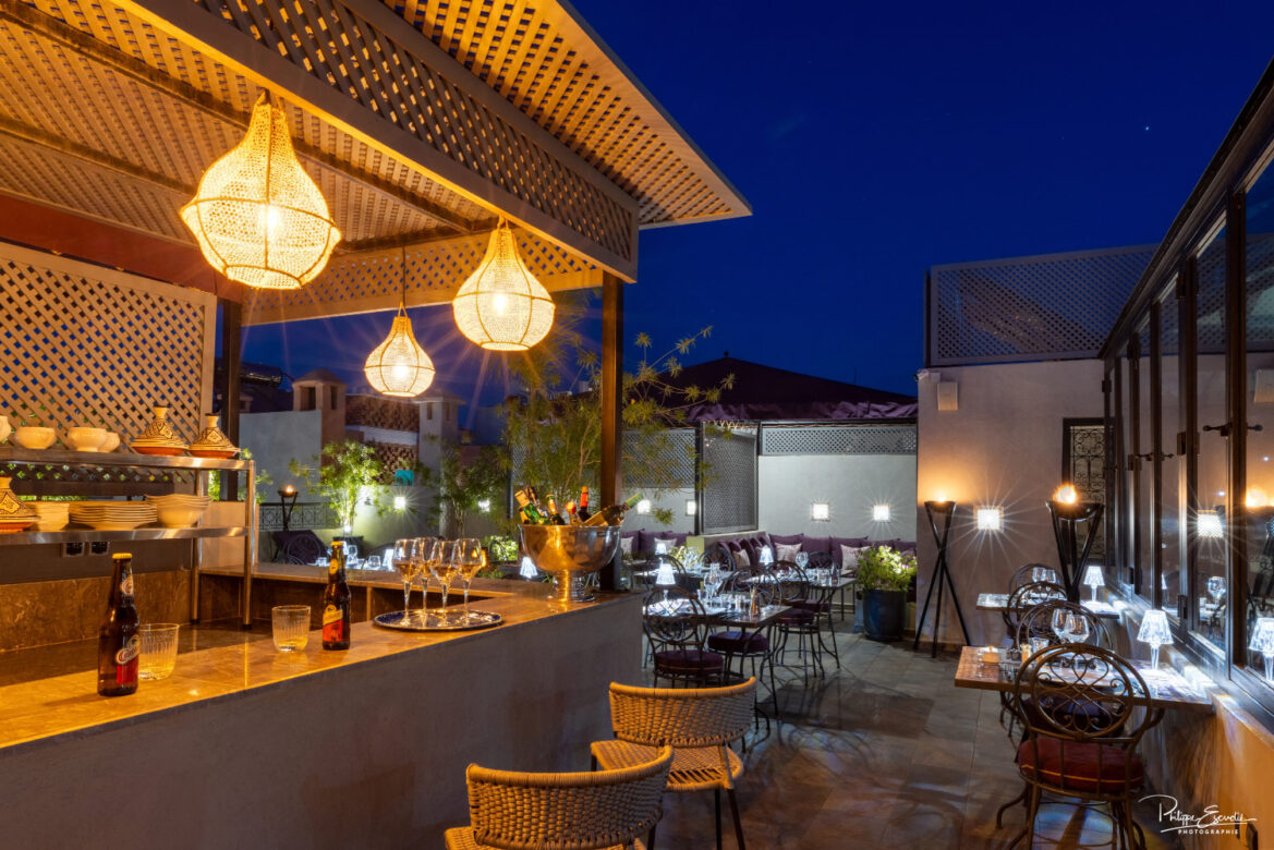 Bar d'une terrasse de riad, atmosphère nuit, avec des boissons sur le comptoir et des tables dressées sur le patio