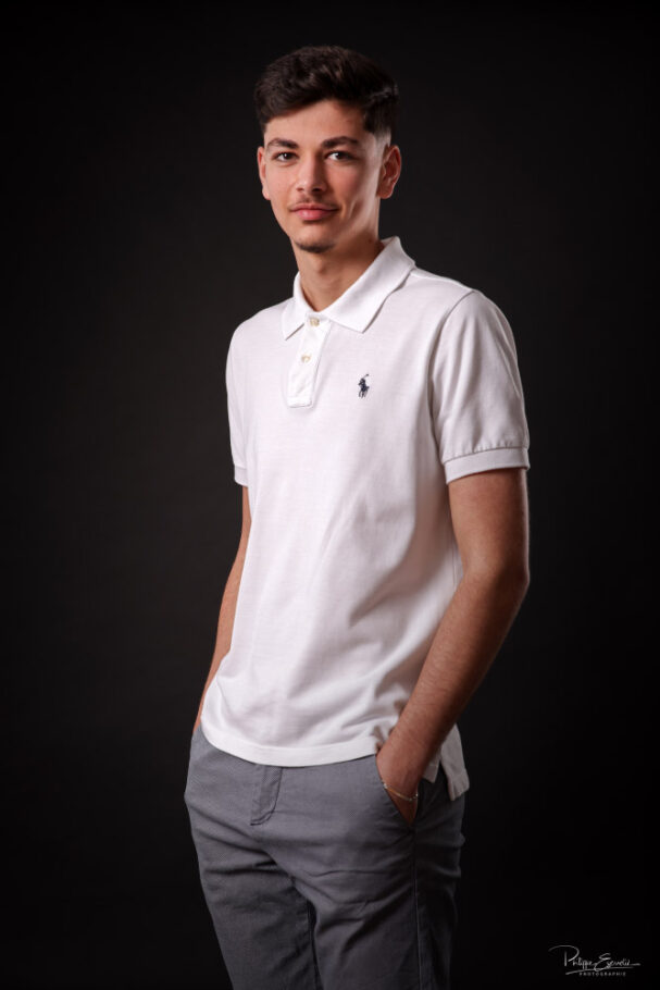 un jeune homme avec un tee shirt blanc les mains dans les poches pose de face sur fond sombre