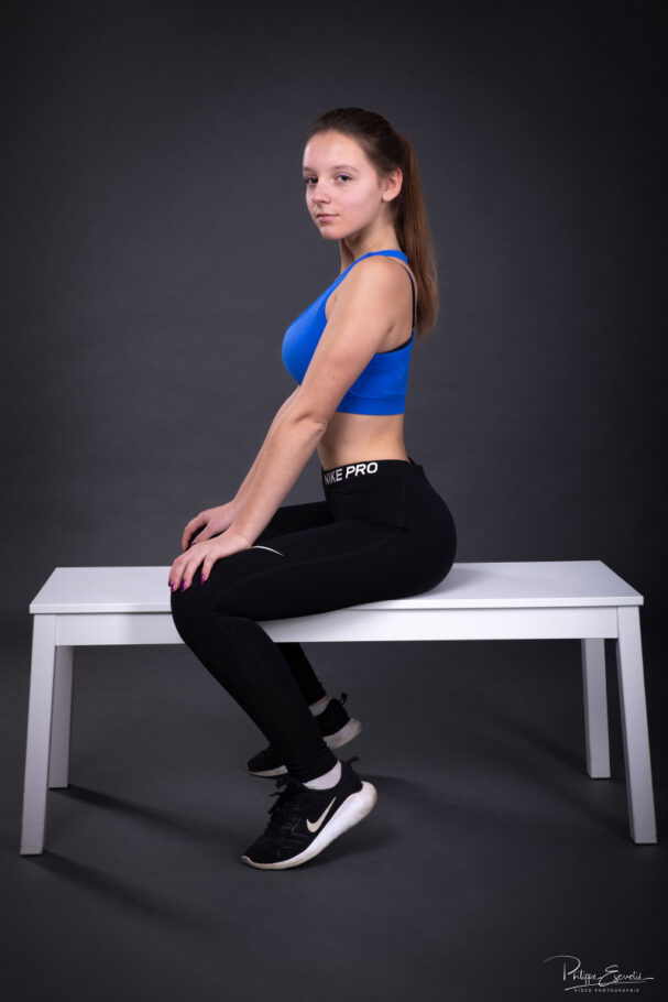 Une jeune femme en débardeur bleu et legging noir est assise sur un banc blanc sur fond sombre de studio
