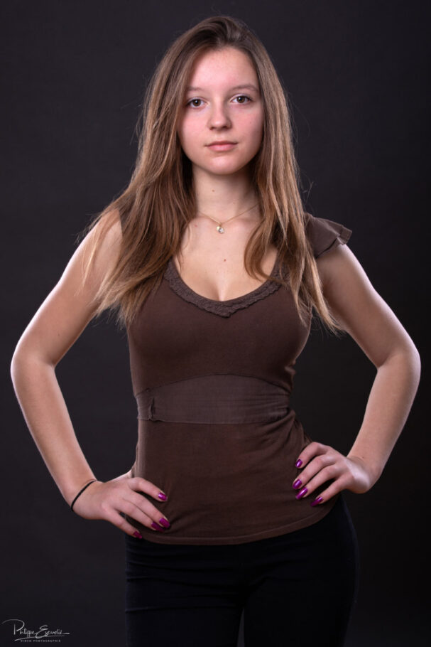 Une jeune femme portant un haut marron les mains sur les hanches pose de face