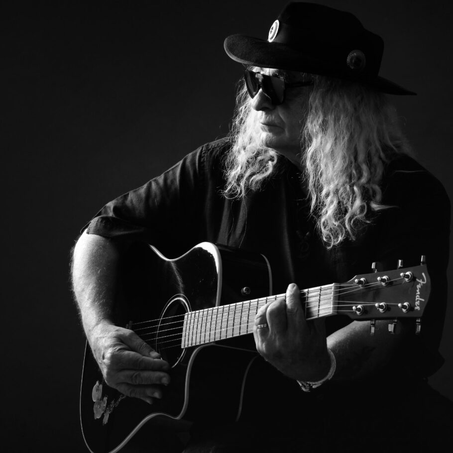Photo noir et blanc en studio fond sombre d'un chanteur assis jouant de la guitare et portant un chapeau et des lunettes de soleil