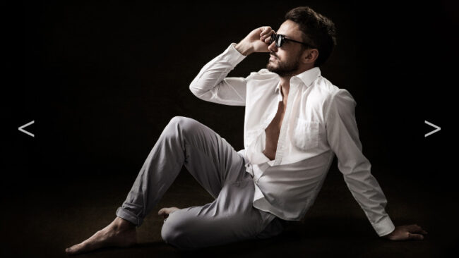 Un homme en chemise blanche et pantalon est assis sur le sol, il porte des lunettes de soleil et affiche une posture détendue et contemplative.