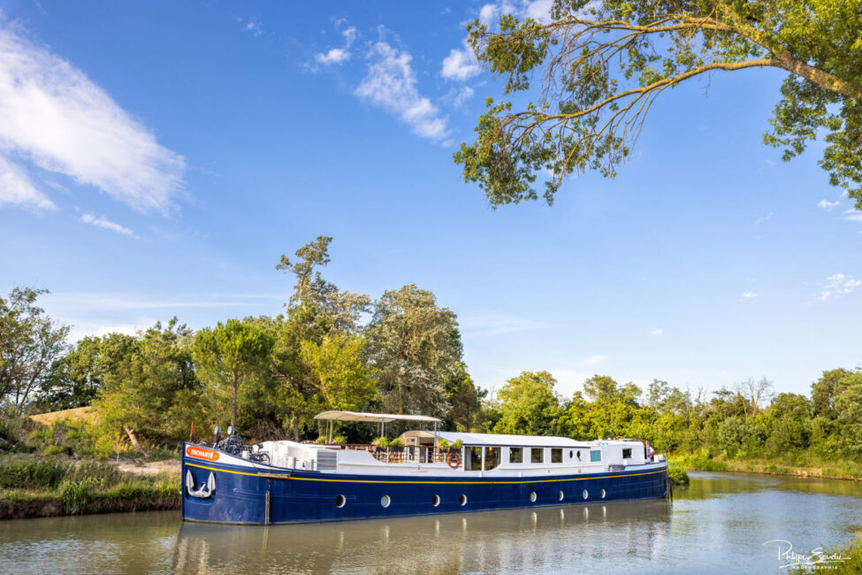 Une grande péniche bleue et blanche navigant sur les eaux vertes d'un canal avec de la végétation en arrière plan