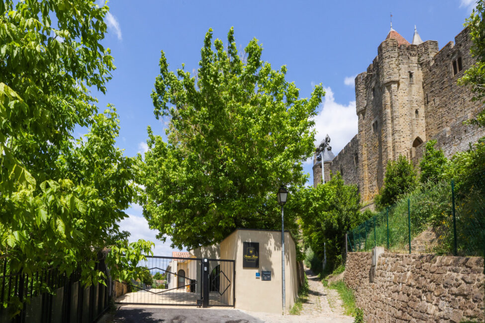 Entrée d'un propriété avec un portail en fer noir, de la végétation autour et sur la droite la Cité de Carcassonne