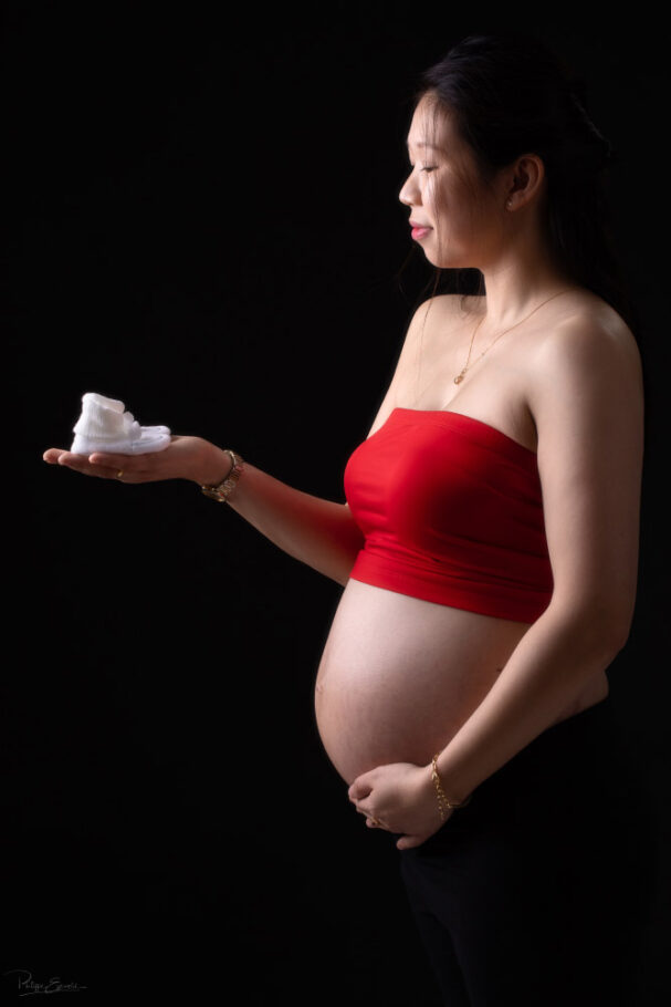 une femme asiatique enceinte de profil porte dans une main des chaussons de bébé