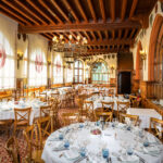 Salle d'un restaurant luxueux avec des tables rondes dressées, de grands lustres fixés au plafond en bois et des tentures aux fenêtres