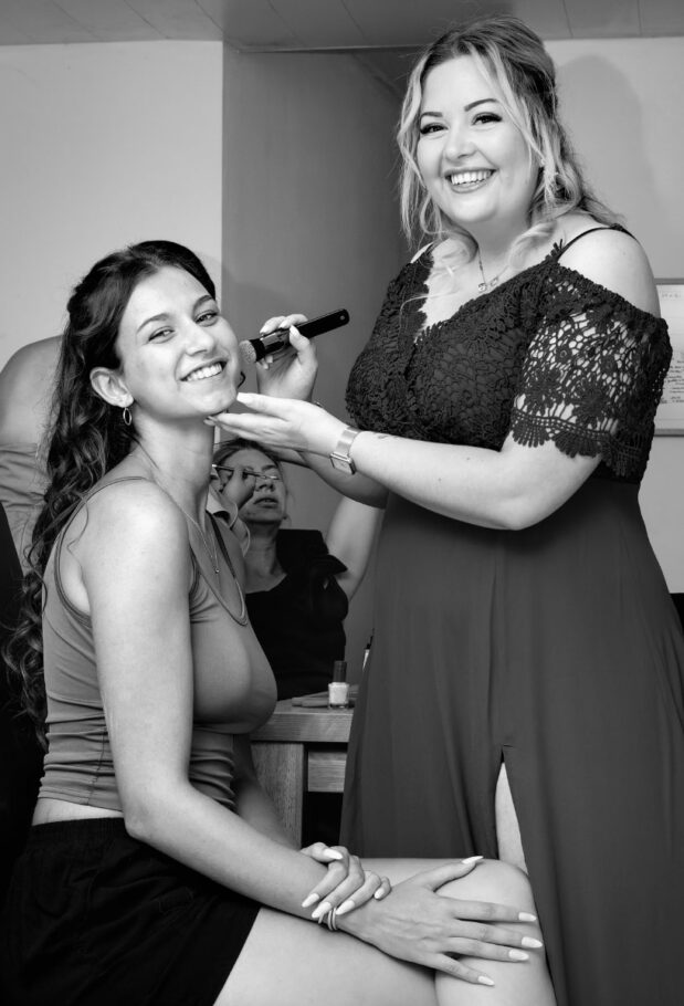 Une fille maquille avec un pinceau une autre fille assise sur une chaise, les deux sourient face objectif