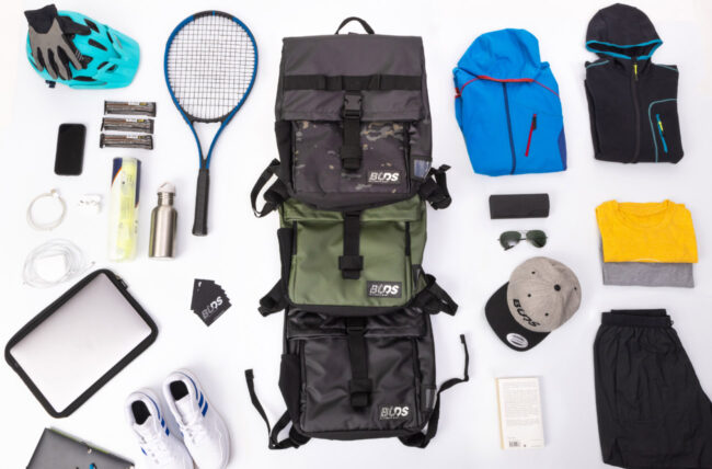 Accessoires de sport posés au sol sur fond blanc, sacs, raquette de tennis, casquette, tee shirt, gants, chaussures, lunettes, vêtements divers