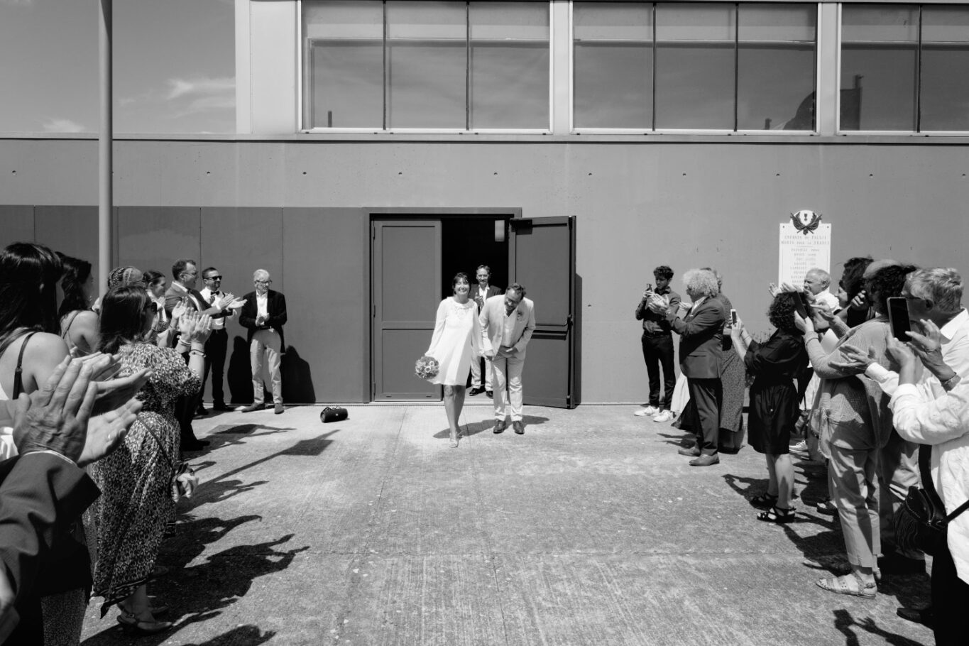 photo noir et blanc, deux mariés sortent d'un bâtiment avec de part et d'autre une rangé de témoins qui les applaudissent