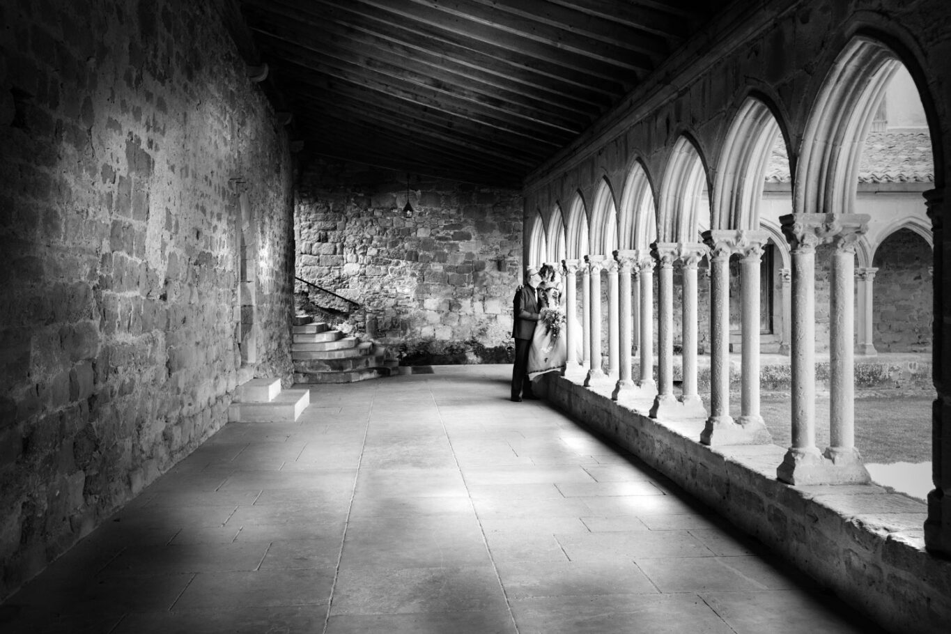 Photo en noir et blanc d'un cloitre avec un couple de mariés qui s'embrasse en arrière plan