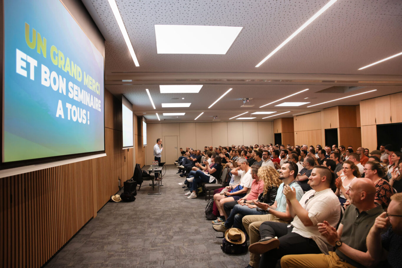 public devant un écran vidéoprojection dans une vaste salle avec en arrière plan un homme qui tient un micro