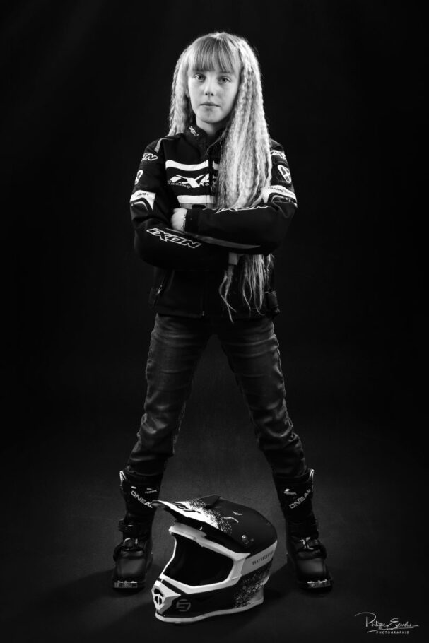 Photo en noir et blanc d'une enfant en tenue de motarde, bras croisés, un casque de moto à ses pied