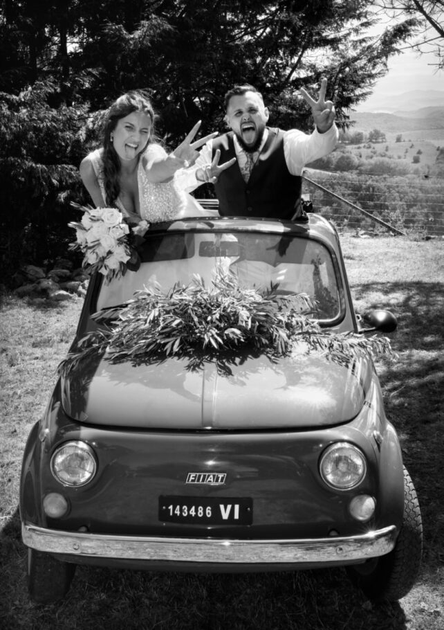 Deux jeunes mariés dans une petite voiture décapotable lèvent les bras et tirent la langue