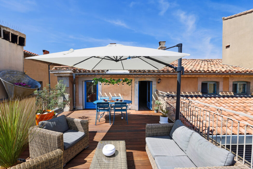 Une grande terrasse en bois avec un salon, un parasol et en arrière plan une table bleue sur laquelle il y a un petit déjeuner