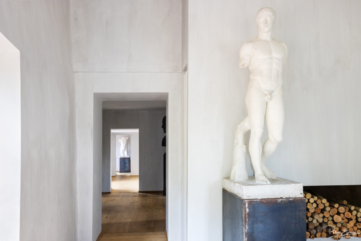 Intérieur d'une maison avec un couloir menant à une statue en marbre, sol en parquet et murs blancs, lumière naturelle - photographie immobilière de prestige