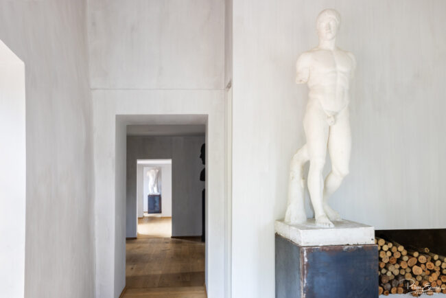 Intérieur d'une maison avec un couloir menant à une statue en marbre, sol en parquet et murs blancs, lumière naturelle - photographie immobilière de prestige
