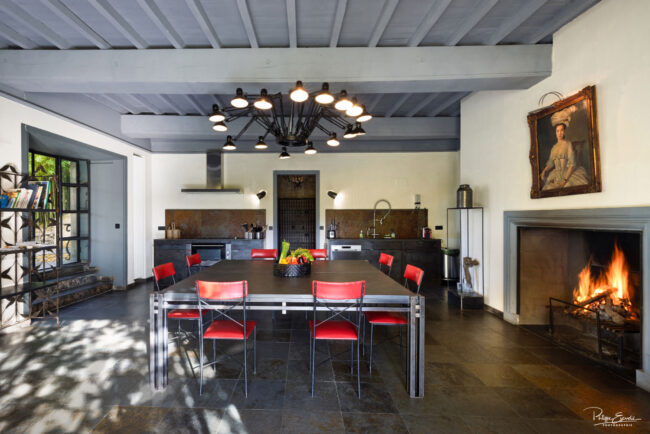 Cuisine spacieuse avec table industrielle, chaises rouges, grande cheminée et éclairage design, dans un style rustique et moderne