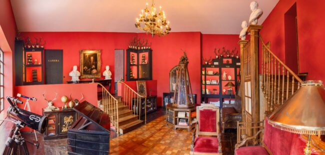 Salle des curiosités au style baroque avec murs rouges, objets anciens, bibliothèque, bustes en marbre et lustre doré