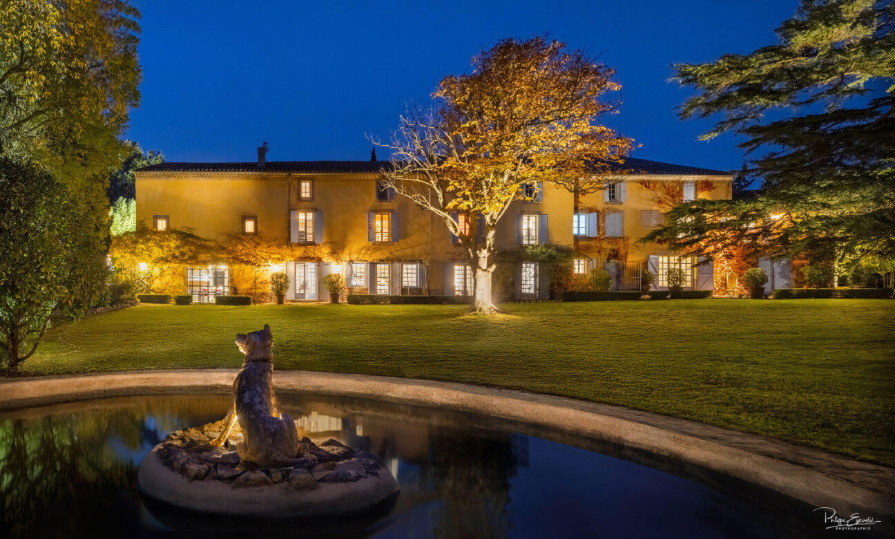 Domaine éclairé de nuit avec parc arboré, fontaine ornée d’une statue de loup et façade en pierre couverte de lierre