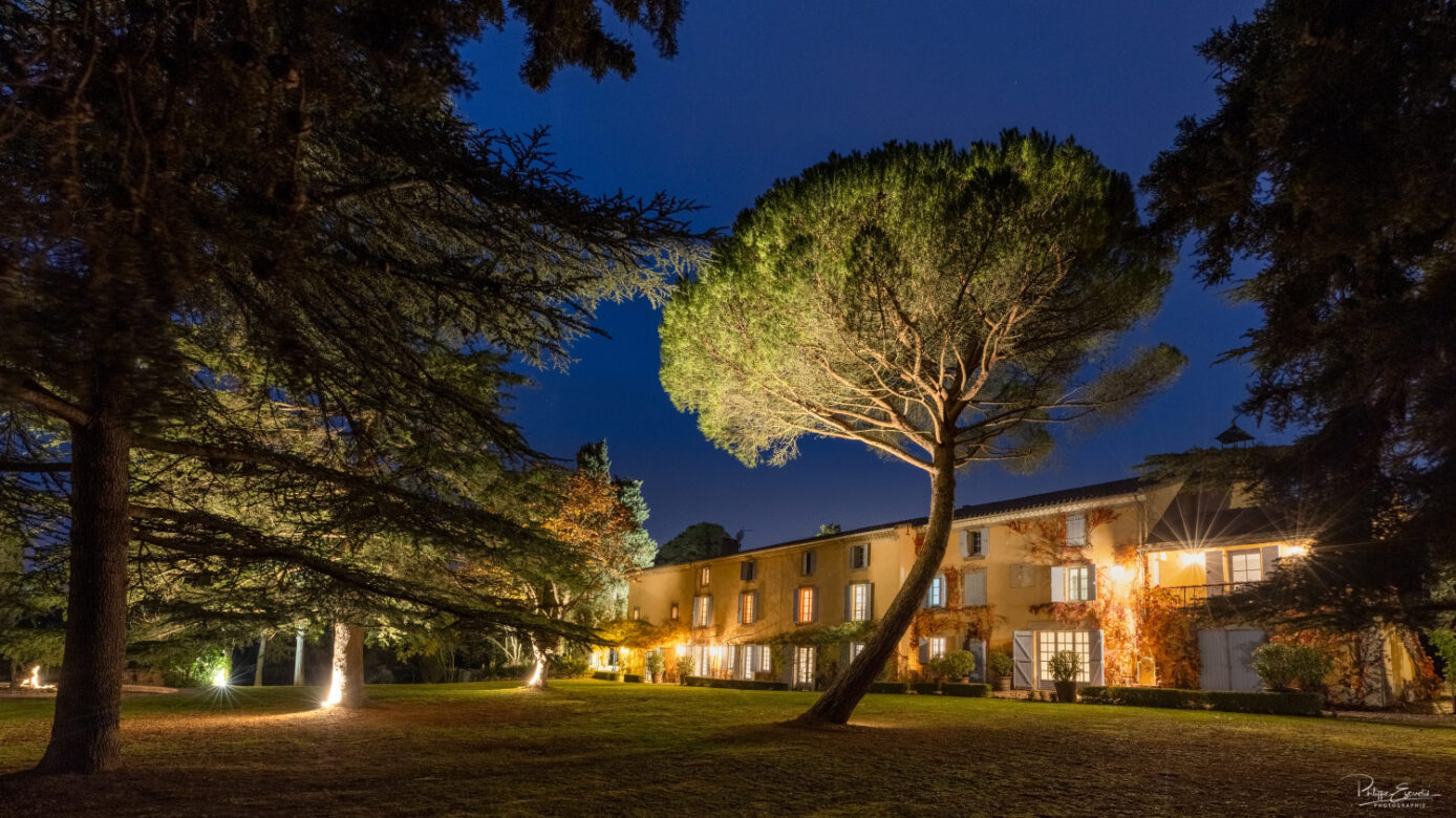 Domaine éclairé de nuit avec façade en pierre, grand parc arboré et ambiance lumineuse raffinée
