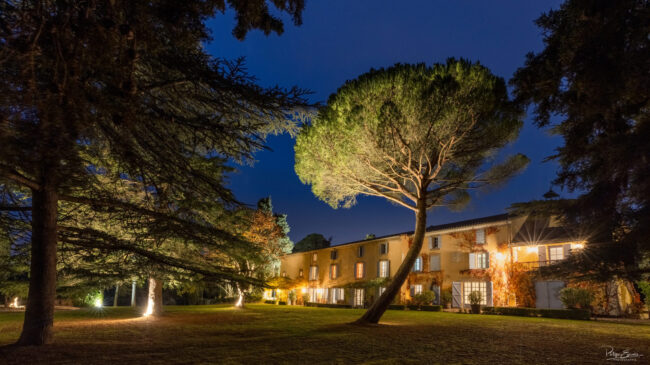 Domaine éclairé de nuit avec façade en pierre, grand parc arboré et ambiance lumineuse raffinée