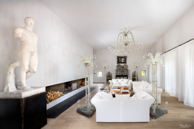 Salon luxueux avec décoration classique et design, grande cheminée moderne, canapé blanc et statue en marbre, éclairé par un lustre en cristal et des luminaires en verre