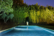 Piscine éclairée de nuit entourée de bambous et oliviers, avec une statue classique en arrière-plan