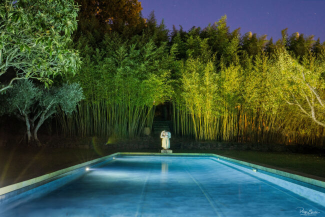 Piscine éclairée de nuit entourée de bambous et oliviers, avec une statue classique en arrière-plan
