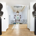 Hall d’entrée élégant avec parquet en bois clair, bustes en bronze représentant le visage de César sur socles noirs, balustrade en fer forgé et lustre suspendu