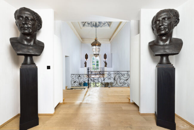 Hall d’entrée élégant avec parquet en bois clair, bustes en bronze représentant le visage de César sur socles noirs, balustrade en fer forgé et lustre suspendu