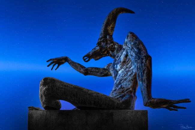 Statue en bronze de créature mythologique avec tête de bouc et posture dynamique, photographiée de nuit sous un ciel étoilé en bleu profond