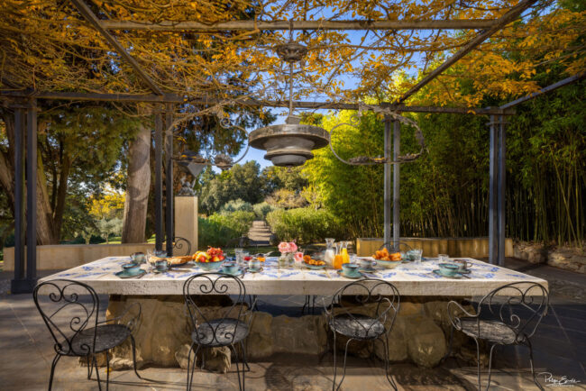 Table en pierre dressée pour le petit-déjeuner sous une pergola ombragée, avec vue sur un jardin luxuriant