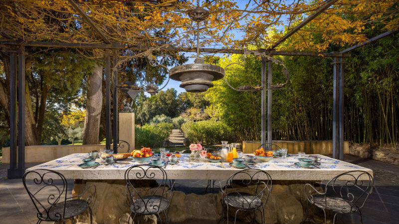 une-table-petit-dejeuner-domaine-bru Table en pierre dressée pour le petit-déjeuner sous une pergola ombragée, avec vue sur un jardin luxuriant