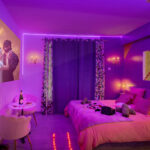 Chambre romantique avec éclairage tamisé violet, projection de film, lit double et accessoires dans une love room