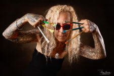 Portrait artistique en studio d'une femme tatouée aux cheveux blonds, un pendentif en forme de tête de mort, tenant des pinceaux colorés devant son visage