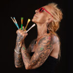 Portrait studio d’une femme tatouée de profil avec pinceaux à la main et des lunettes rouge – photo pour book