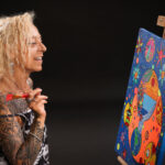 Portrait en studio d’une femme tatouée souriante tenant un pinceau devant une toile colorée sur chevalet –pour book d’artiste