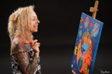 Portrait en studio d’une femme tatouée souriante tenant un pinceau devant une toile colorée sur chevalet –pour book d’artiste