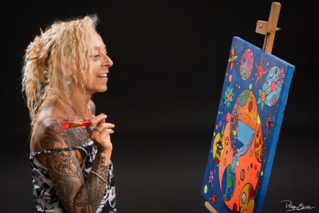 Photographe Portrait : Focus sur Cristelle Bernardinello, artiste peintre 2 Portrait en studio d’une femme tatouée souriante tenant un pinceau devant une toile colorée sur chevalet –pour book d’artiste