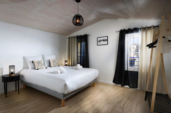 Chambre lumineuse sous combles avec lit double, rideaux bicolores, deux tables de chevet et plafond en bois clair