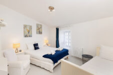 Chambre blanche décorée de tableaux de cerfs, lit double avec plaid bleu marine et fauteuil d’appoint.