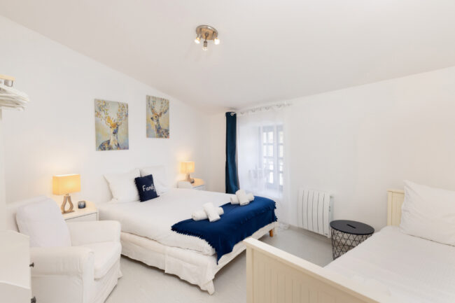 Chambre blanche décorée de tableaux de cerfs, lit double avec plaid bleu marine et fauteuil d’appoint.
