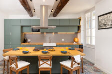 Cuisine équipée moderne avec grands placards verts, îlot central en bois et vaisselle dressée pour le petit déjeuner