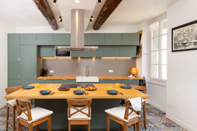 Cuisine équipée moderne avec grands placards verts, îlot central en bois et vaisselle dressée pour le petit déjeuner