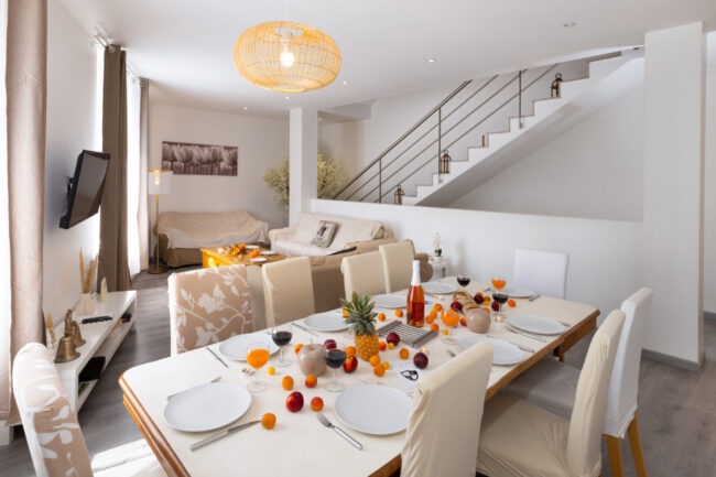 Salon lumineux avec coin repas décoré de fruits, canapés clairs, escalier et luminaires suspendus