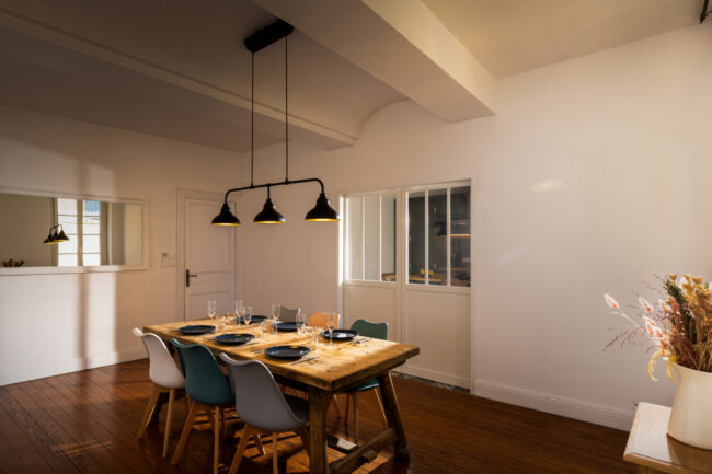 Salle à manger avec grande table en bois, décoration florale, parquet en bois foncé et lumière tamisée.