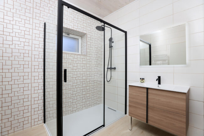 Salle de bain moderne avec douche à l’italienne, paroi vitrée noire, carrelage géométrique et meuble en bois clair.