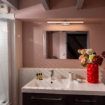 Salle de bain rose avec vasque blanche, miroir mural, bouquet de fleurs et douche avec paroi striée.