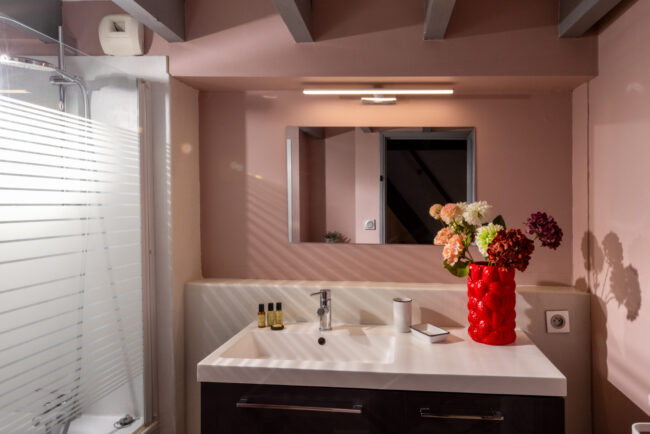 Salle de bain rose avec vasque blanche, miroir mural, bouquet de fleurs et douche avec paroi striée.